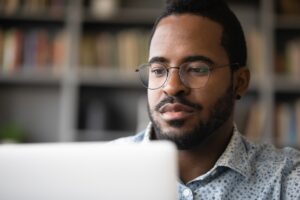 African American man sitting in front of bookshelf looking at laptop.