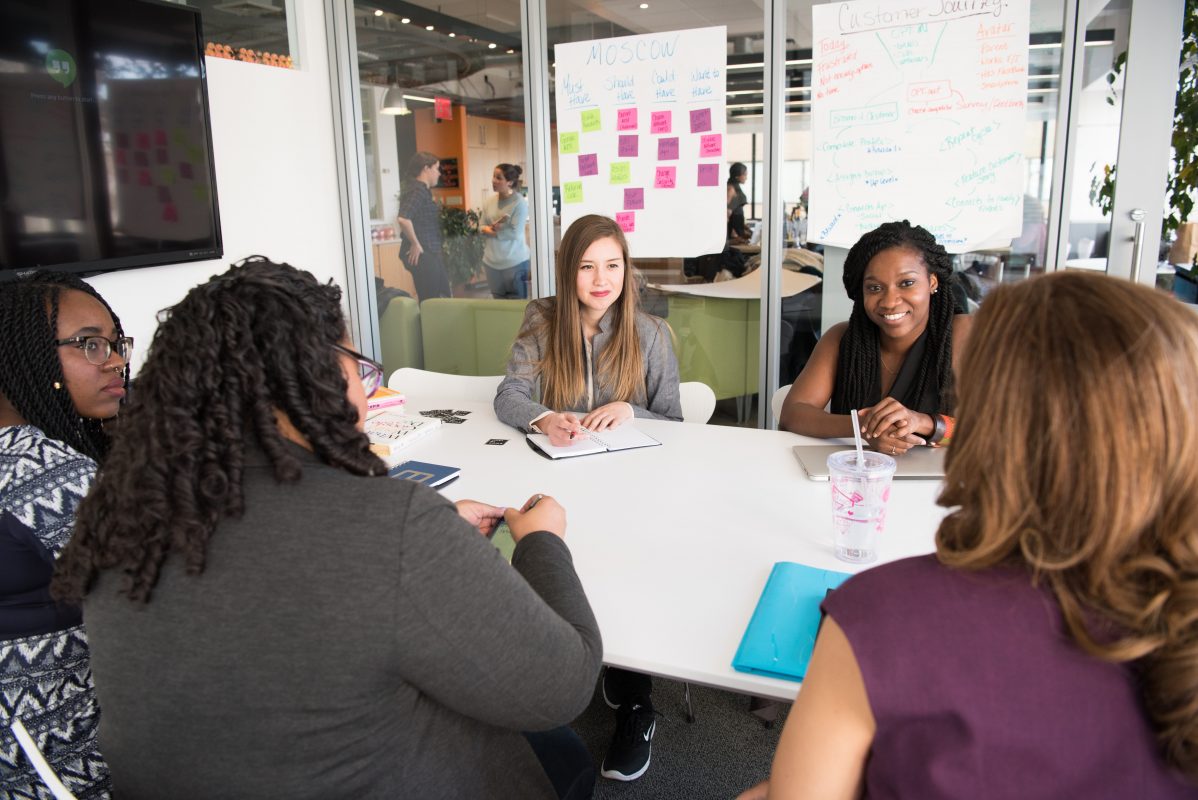 A group of women engaged in conversation around a meeting table in front of a wall with post it notes on it.