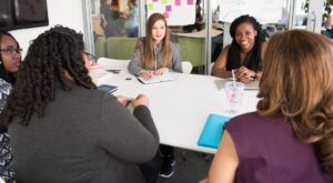 A group of women engaged in conversation around a meeting table in front of a wall with post it notes on it.