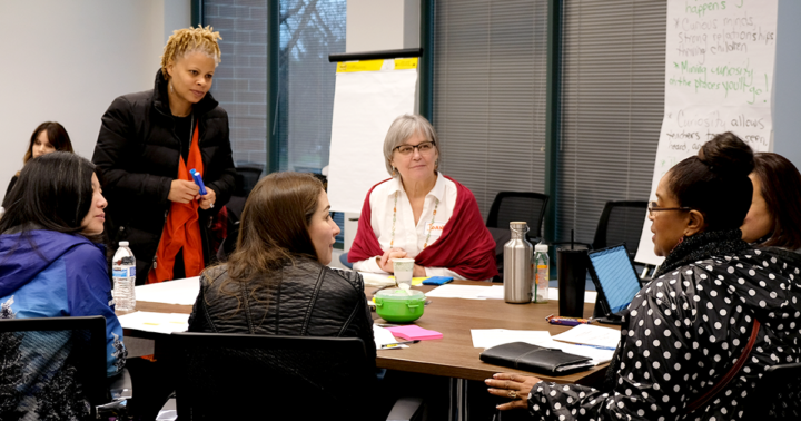A group of educators seated around a table listening to one person talking as another person stands prepared to take notes on a flip chart nearby.