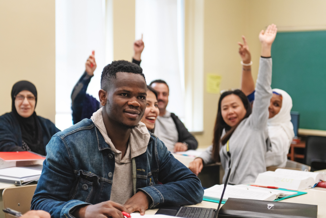 Class of adult students, with one student featured in the forefront and other students seated in a group behind him raising their hands. All look happy, engaged.
