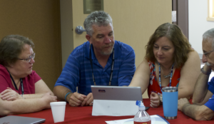 A group of TCU faculty and a faculty mentor seated around a table looking together at a laptop.